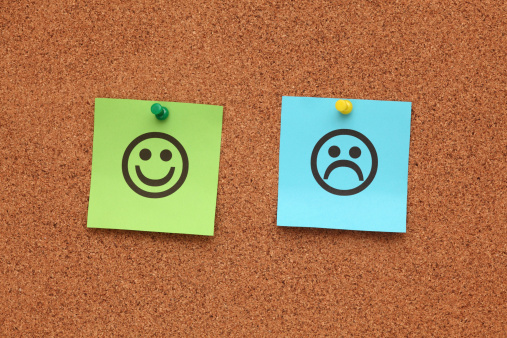 Two sticky notes posted to a corkboard; one is green and has a smiley face drawn on, the other is blue and has a frowny face drawn on.