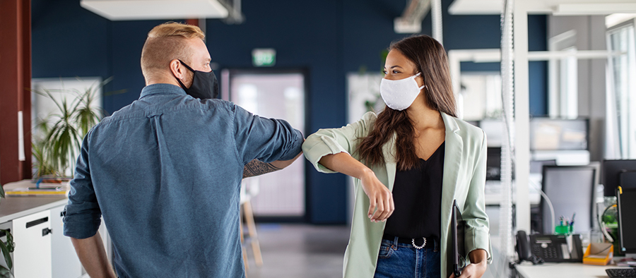 Two people in wearing business-casual attire and fabric face masks bump elbows to greet each other.