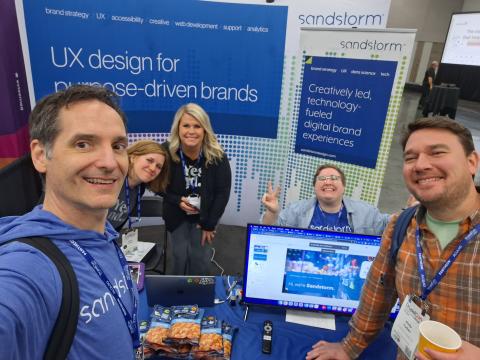Five members of the Sandstorm Drupal web agency are gathered at their booth at Drupalcon. The signage says, “UX design for purpose-driven brands. Creatively led, technology-fueled digital brand experiences.” They are smiling at the camera.  
