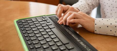 A blind woman uses a computer with a Braille display and a computer keyboard. Inclusive device.