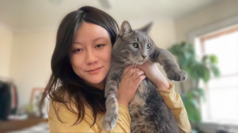 Vivian Young is a female with long brown hair, brown eyes, wearing a yellow sweater, smiling at the camera holding her grey cat