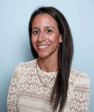 A professional headshot of a woman with long, straight dark hair, wearing a ivory crochet blouse and layered gold necklaces. She is smiling warmly against a light blue background.